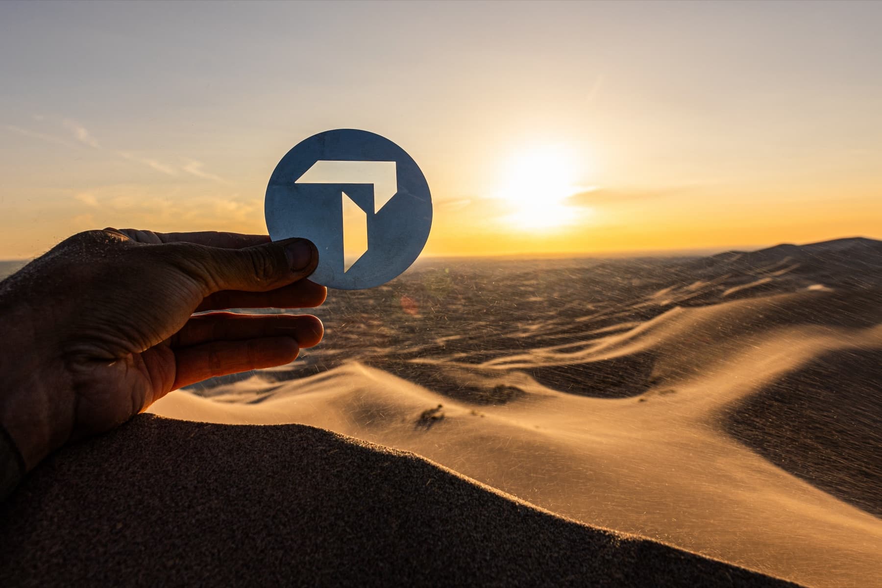 Ruka drží kovový symbol Travelport na pozadí písečných dun pouště Gobi při západu slunce