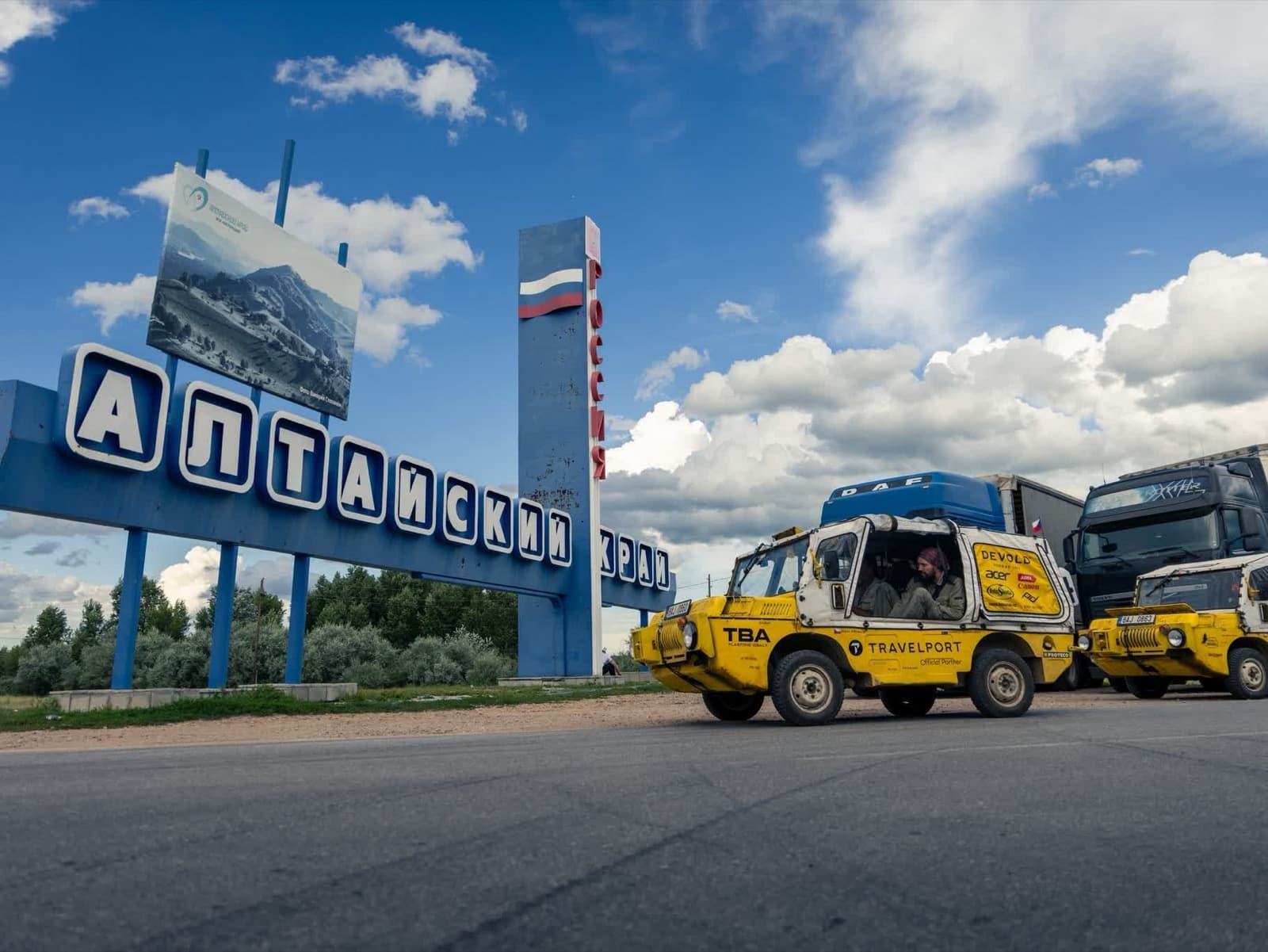 Žlutý expediční vůz s nápisem Travelport projíždí kolem cedule Altajského kraje na hranici s Ruskem