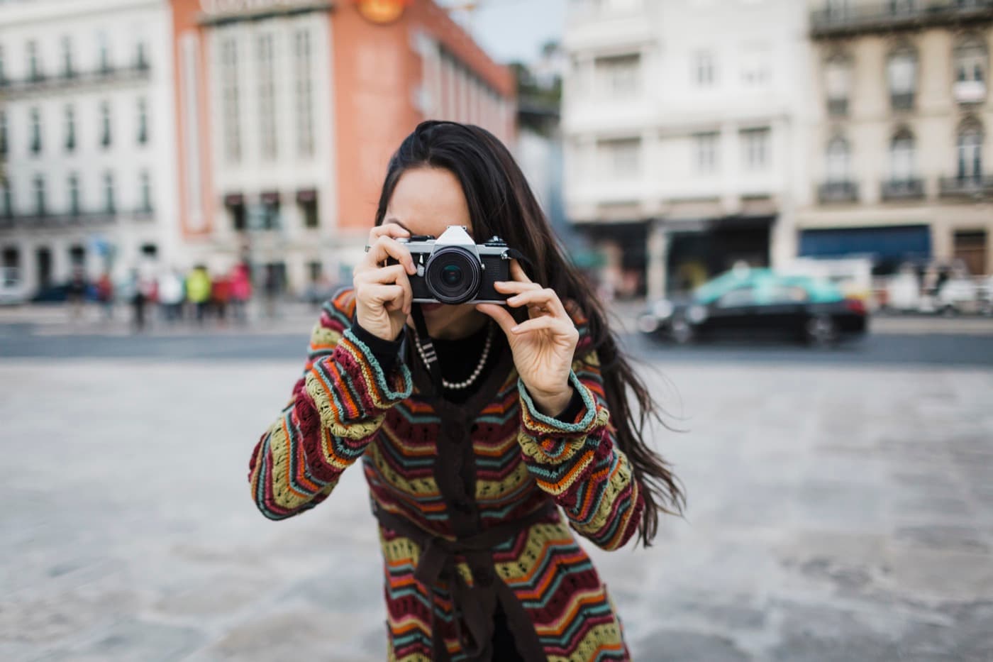 Fotografka v pestrém svetru míří objektivem analogového fotoaparátu přímo na diváka
