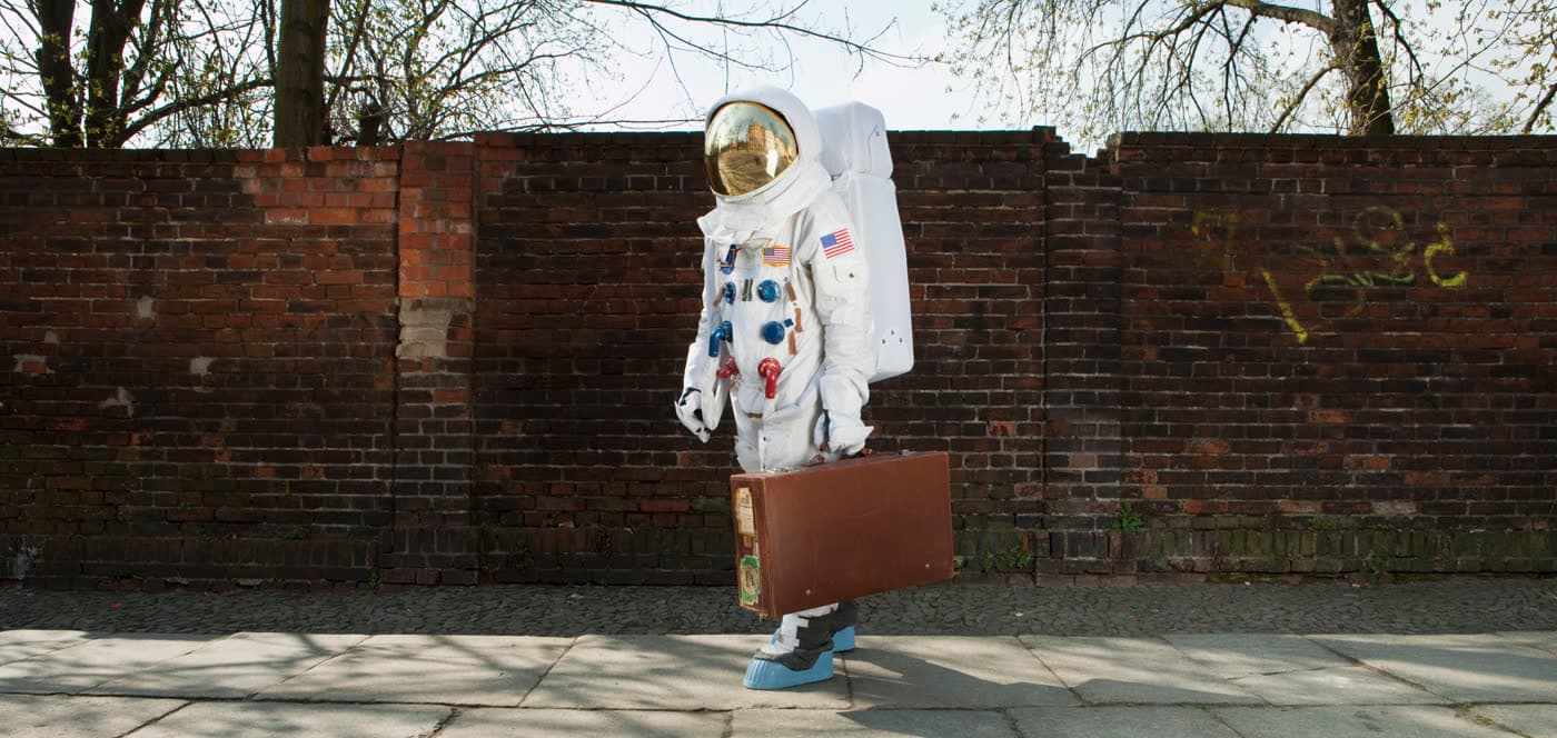 Astronaut s koženým kufrem v ruce čeká na chodníku u cihlové zdi