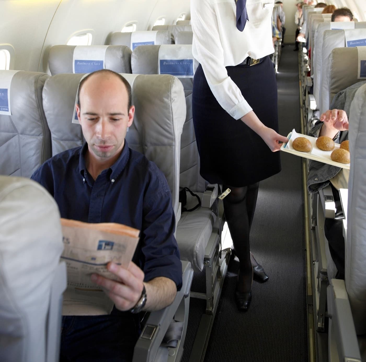 Letuška nabízí pečivo cestujícím business class, muž si čte noviny